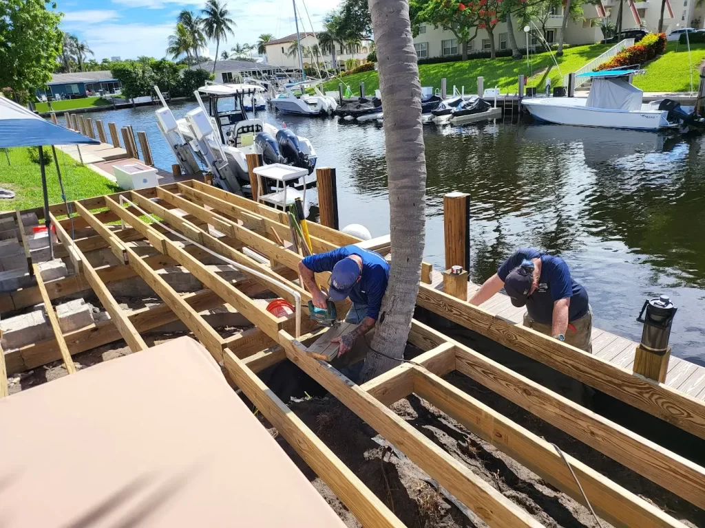 The image depicts workers constructing or repairing a seawall along a waterfront, with wooden beams and supports in place. The scene includes a canal with boats, palm trees, and residential buildings in the background under a clear blue sky.